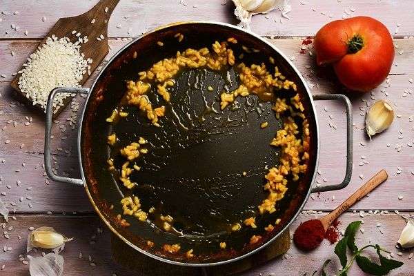 paellera con restos de arroz amarillo sobre mesa de madera con cuchara de palo con arroz crudo, tomates y ajo