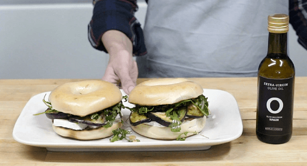 Mano mostrando plato blanco con bagels rellenos de berenjena, miel y queso brie con una botella de aceite de oliva al lado