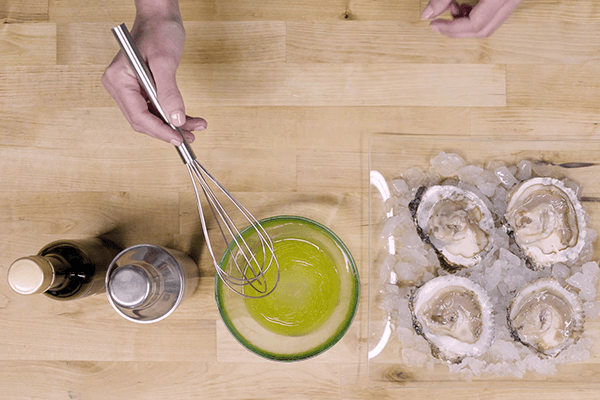 Menos batiendo con zumo de limón y aceite de oliva en recipiente de crista sobre mesa de madera. Al lado, plato con hielo y ostras.