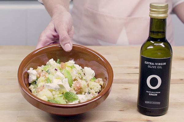 Mano mostrando recipiente de barro con couscous de verduras y botella de aceite de oliva virgen extra