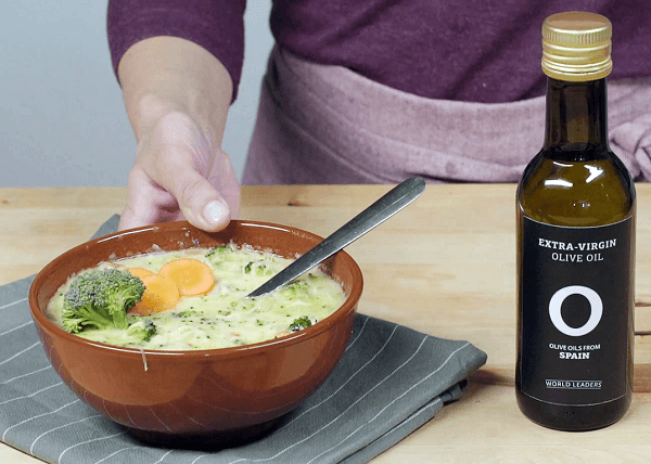 Mano mostrando cuenco de barro con crema de brócoli, al lado una botella de aceite de oliva. 