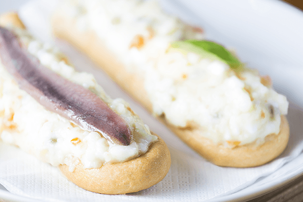 Pan con ensaladilla rusa y anchoa sobre servilleta blanca en un plato blanco. 