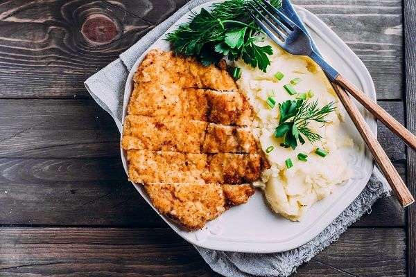 FIlete empanado acompañado de puré de patatas en plato blanco con cubiertos sobre madera