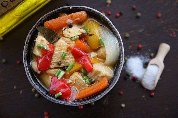 cazuela con pollo con verduras sobre tabla negra con cuchada de palo con sal y pimienta