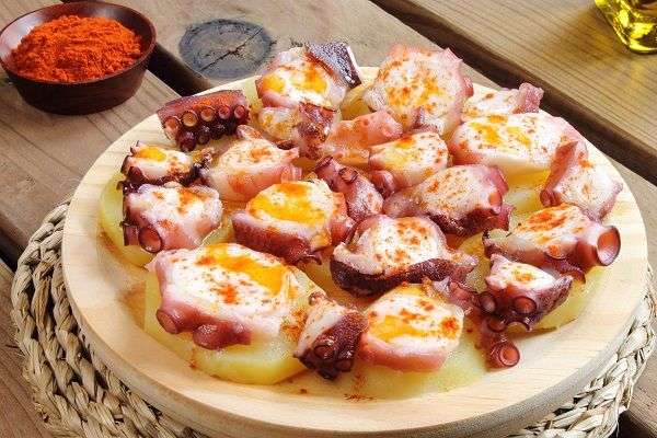 Pulpo a la gallega con pimentón y patata en tabla redonda sobre salvamanteles cobre mesa de madera con bol marrón con pimentón y botella de aceite de oliva