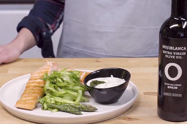 Plato blanco con salmón a la parrilla con espárragos y salsa de yogurt en un recipiente negro decorado con un espárrago verde al lado de botella de aceite de oliva sobre mesa de madera. Al fondo persona con delantal. 