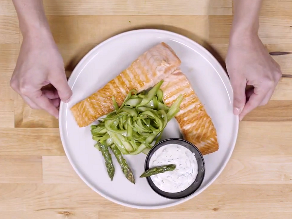 Manos mostrando un plato blanco con salmón a la parrilla con espárragos y salsa de yogurt en un recipiente negro decorado con un espárrago verde.