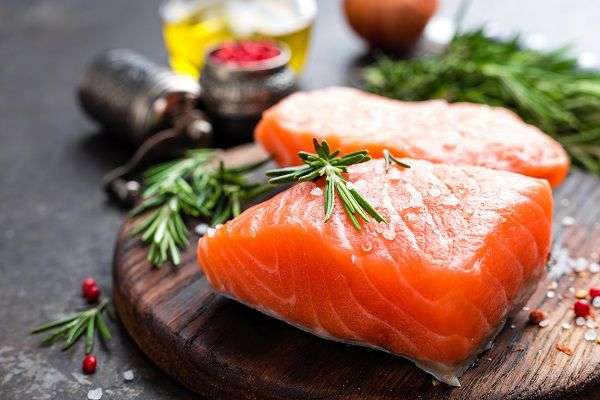 Salmón sobre tabla de madera decorado con ramas verdes. Al fondo recipiente con aceite de oliva. 