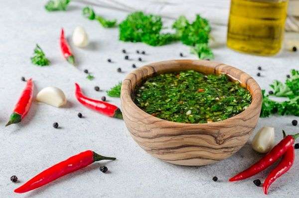 Salsa chimichurri en recipiente de madera sobre mesa blanca con adornos de hierbas aromáticas, una botella de aceite de oliva y semillas de pimienta