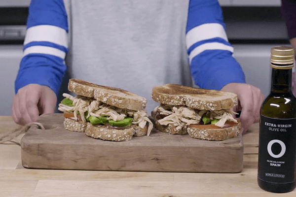 Brazos de niño mostrando sándwich de pollo mechado con paprika y aguacate sobre tabla de madera en mesa de madera con una botella de aceite de oliva