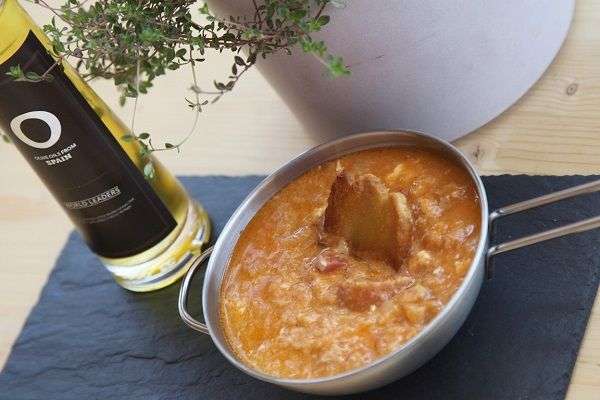 Sopa de ajo en cazuela de metal con asas sobre plato de pizarra y botella de aceite de oliva. Al fondo planta en jarrón blanco. 