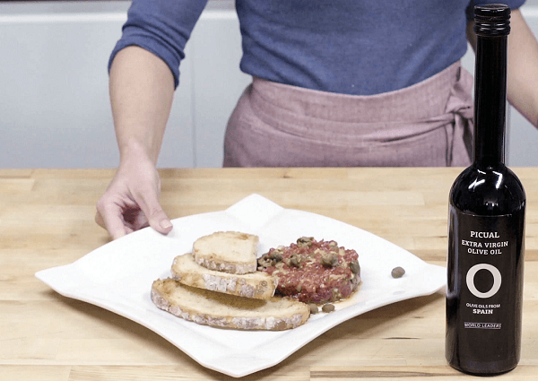 Mano mostrando plato blanco con steak tartar con pan al lado de una botella de aceite de oliva sobre mesa de madera