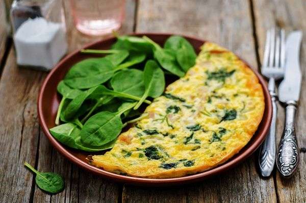 Tortilla de espinacas con espinacas crudas en plato marrón sobre mesa de madera con cubiertos de plata, salero y vaso de agua