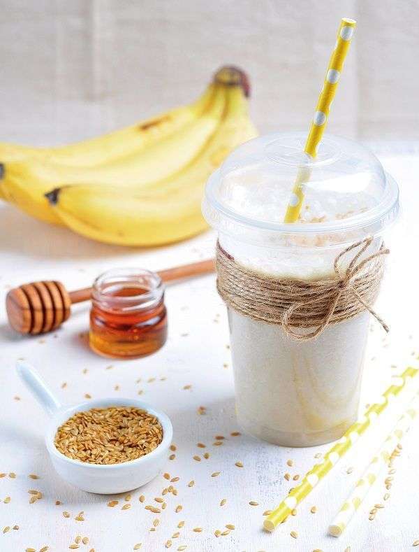 Plátanos, tarro con miel, batido de color blanco en vaso de plástico con lazo y pajita, y bol con semillas sobre mesa blanca 